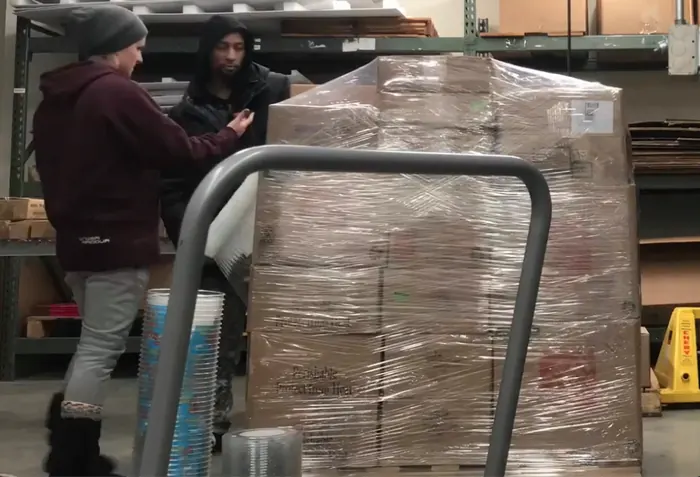 Two workers shrink-wrapping a loaded pallet at the packing area, with warehouse racking behind them.