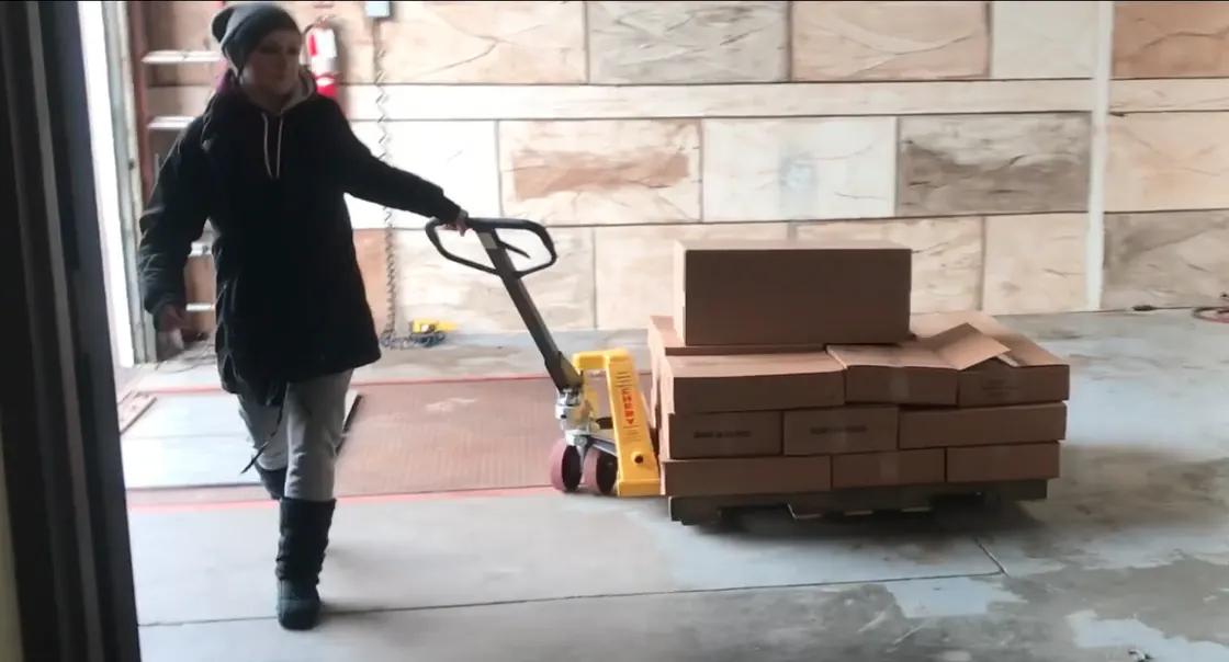 A worker pulls a pallet of stacked cartons in through the warehouse loading door on a pallet jack.