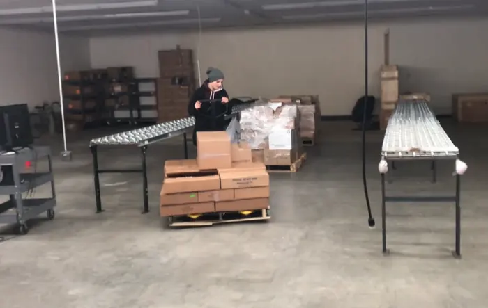 A worker moves a pallet of cartons across the warehouse floor with a pallet jack, beside roller conveyor tables and shrink-wrapped pallets.